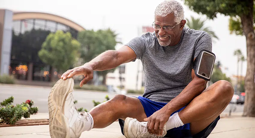 Smiling senior African American man stretching before exercise, wearing gray t-shirt and blue shorts, with smartphone in armband, demonstrating active and healthy lifestyle for older adults