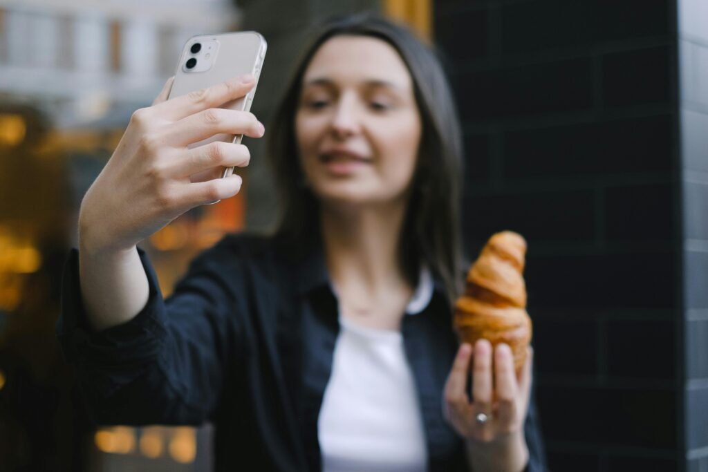 pexels-photo-12679986-12679986 A woman takes a selfie while holding a croissant in an urban outdoor setting.