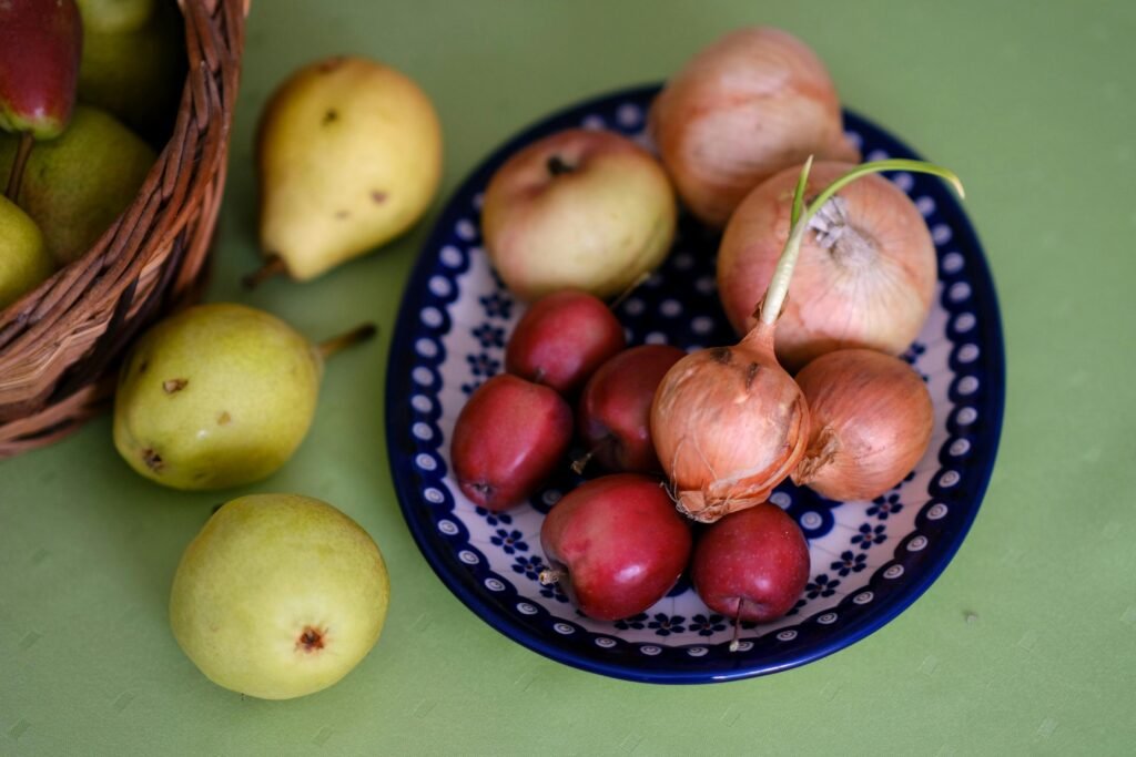 pexels-photo-33818433-33818433 Close-up of pears, onions, and crab apples in a blue patterned bowl on a green surface.
