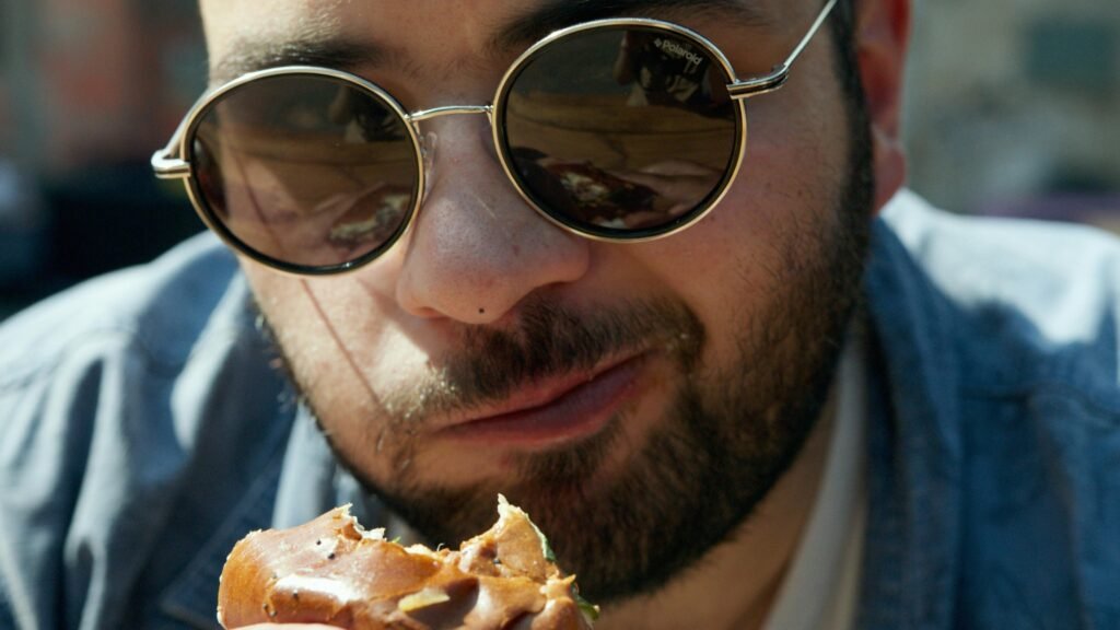 pexels-photo-4728859-4728859 Portrait of a young man wearing sunglasses enjoying a delicious burger outside on a sunny day.