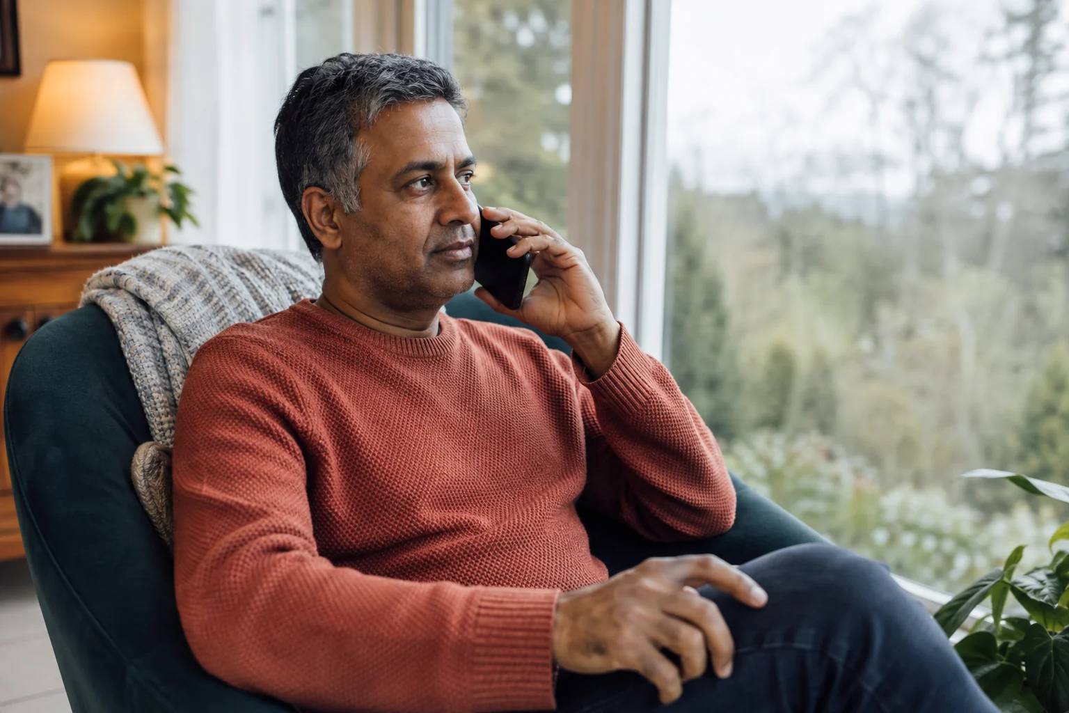 An adult man listening on a phone in a teal armchair beside a bright home window