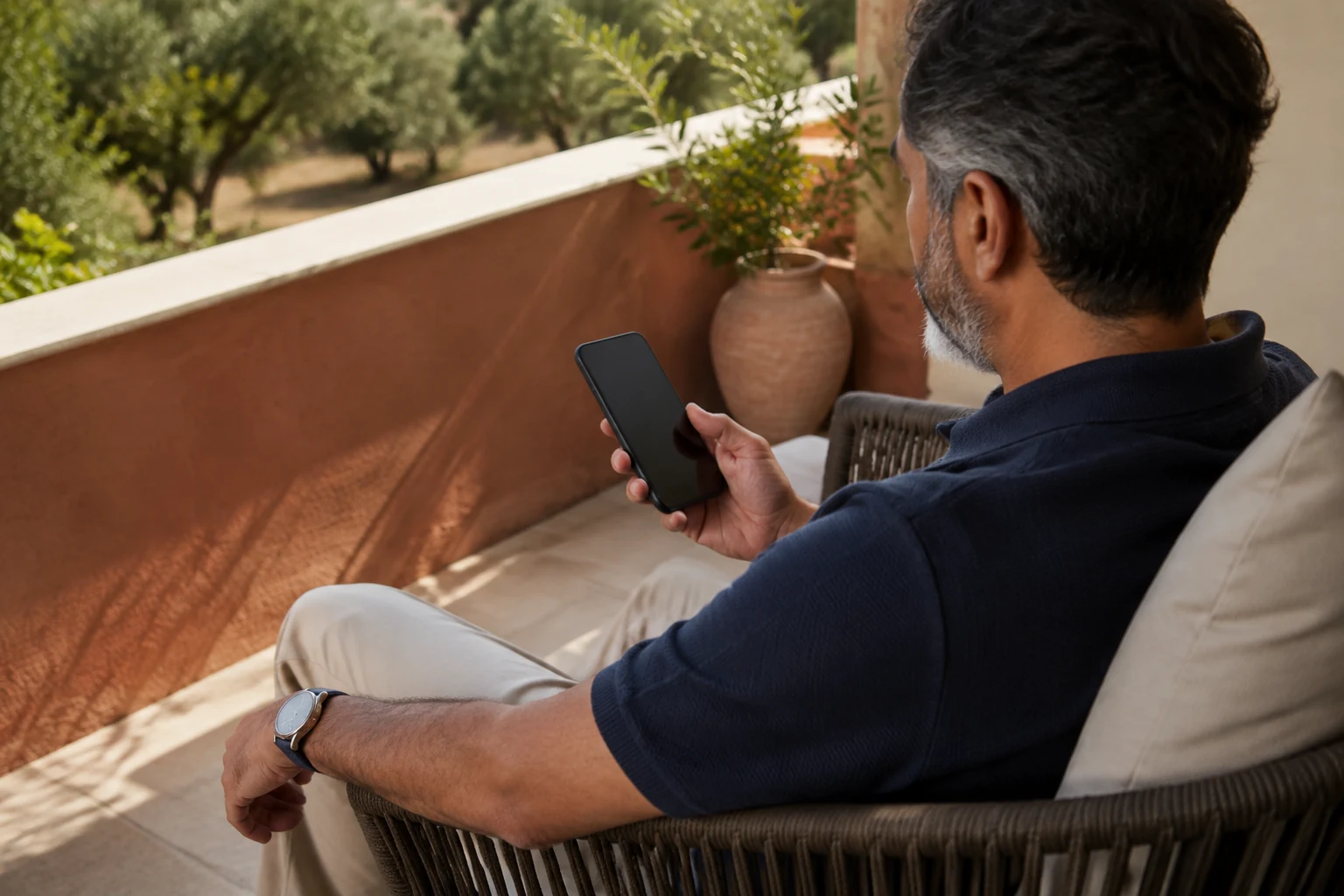 Man in a navy polo checking a smartphone on a sunlit terrace