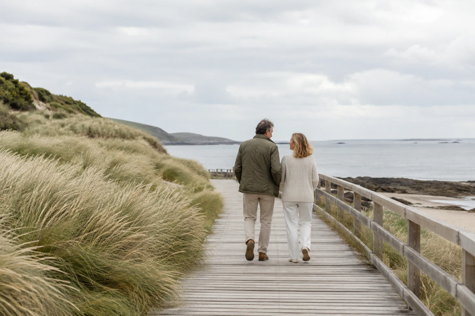 An adult couple walking together along a coastal boardwalk beside dune grasses