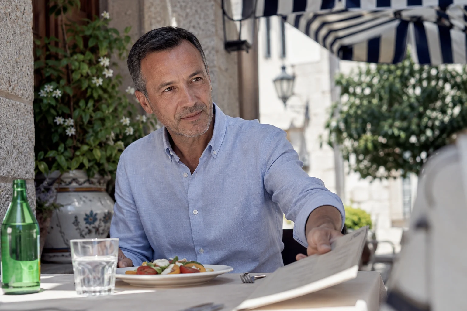 A man setting down a menu at an outdoor bistro beside his lunch and a glass of water