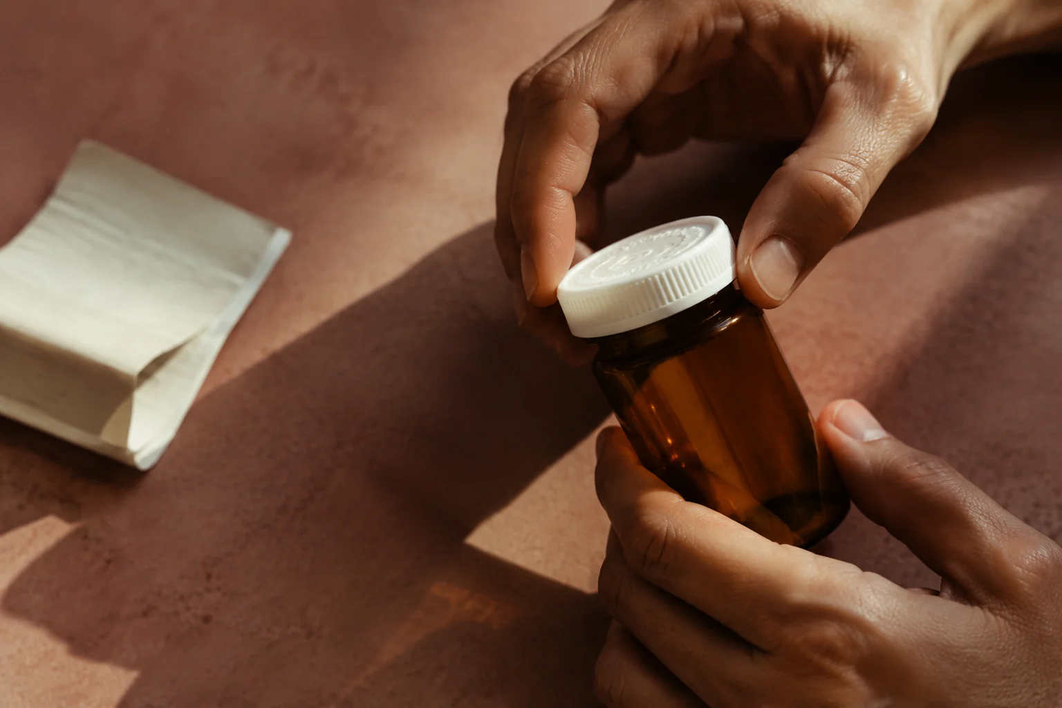Hands opening an unlabelled amber medicine vial on a warm terracotta surface
